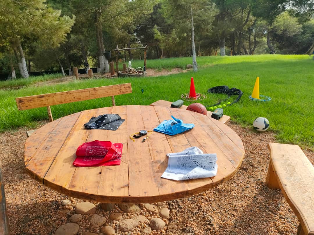 Wooden round table with colorful bandanas and sports equipment on a grassy area with activity cones, surrounded by pine trees
